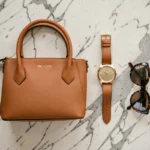 Accessoires de la marque Michael Kors disposés en flat lay sur marbre : sac à main en cuir caramel, montre dorée et lunettes de soleil écaille