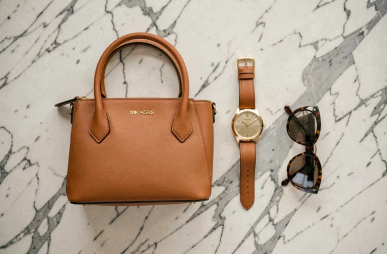 Accessoires de la marque Michael Kors disposés en flat lay sur marbre : sac à main en cuir caramel, montre dorée et lunettes de soleil écaille